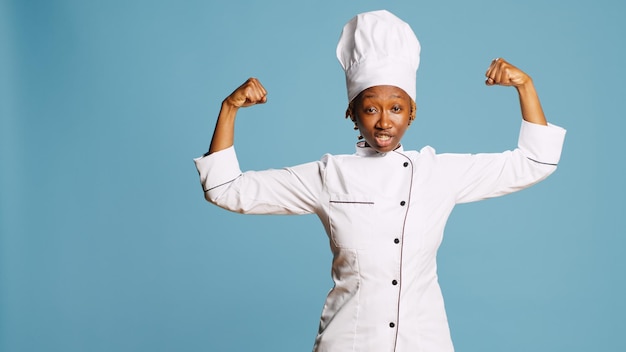 Una mujer poderosa cocina flexionando los músculos del brazo en la cámara, mostrando poder y fuerza corporal en el estudio. Confiado chef feliz en delantal de cocina siendo bueno en la industria de la gastronomía y la cocina.