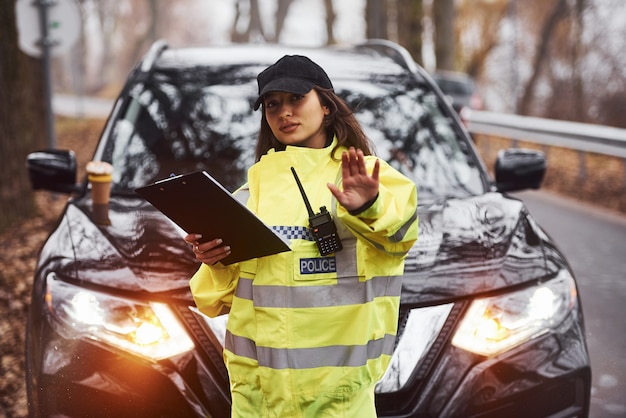 Mujer policía en uniforme verde de pie con transmisor de radio y bloc de notas contra el coche.