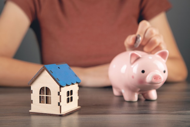 Una mujer pone una moneda en una alcancía y al lado de una casa.