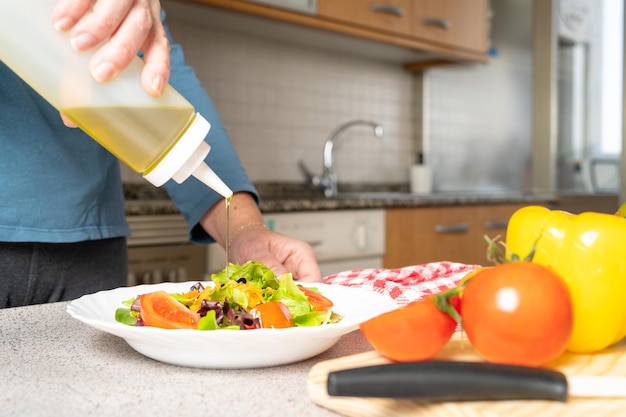 Mujer poniendo aceite de oliva en una ensalada fresca de tomates, lechuga y zanahorias en la cocina de casa