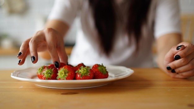 Foto mujer poniendo fresas en un plato cerca de las manos de las mujeres poniendo bayas frescas en un plato blanco en la mesa de la cocina