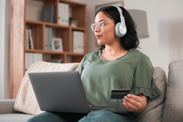 Mujer portátil y tarjeta de crédito con auriculares y compras en línea en el sofá música pago de suscripción y fintech Cliente femenina en el hogar pensando en finanzas con comercio electrónico y membresía de podcast
