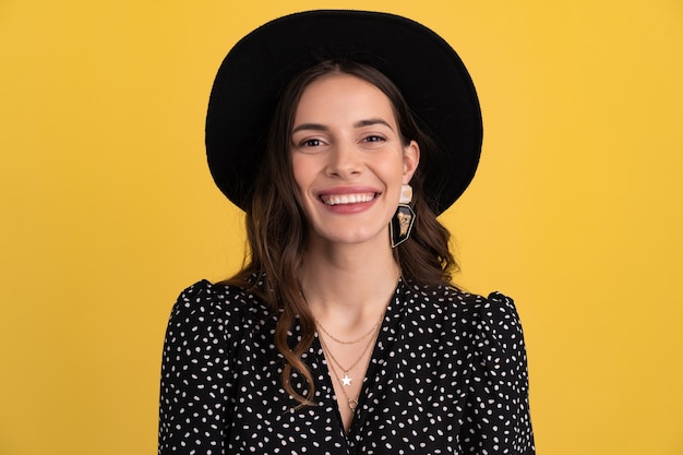 Mujer posando aislada en amarillo con vestido negro punteado y sombrero negro con estilo boho