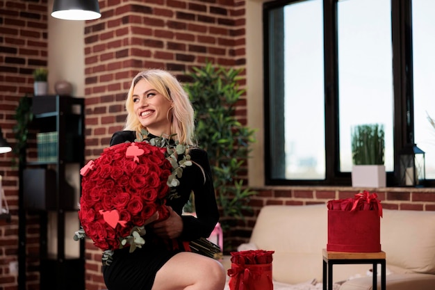 Mujer posando con ramo de rosas rojas de lujo, riendo, sentada en un taburete de bar en la sala de estar, plano medio. Hermosa chica rubia con regalo caro de San Valentín, cita romántica.