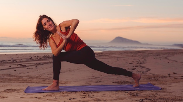 Mujer en pose de yoga en la alfombra en la arena de la playa al amanecer