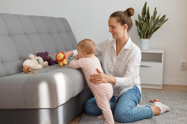 Mujer positiva con camisa blanca y jeans sentada en el suelo con su pequeña hija parada cerca del sofá y jugando con juguetes madre abrazando a su hijo y sonriendo