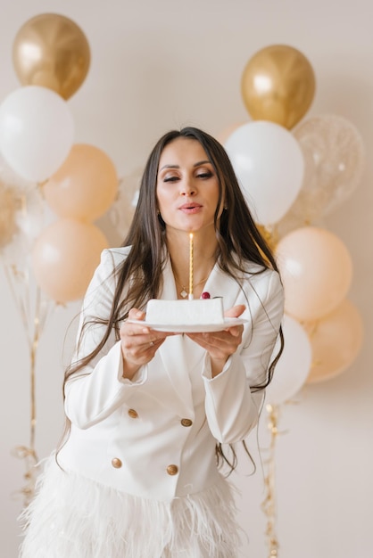 Mujer positiva celebrando el aniversario vestida con un elegante vestido de plumas que sopla una vela en un pastel de cumpleaños alrededor de globos inflados en casa