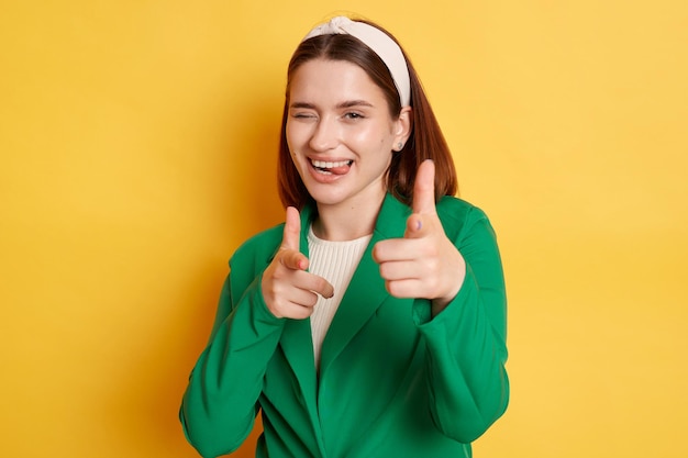 Mujer positiva coqueteando con chaqueta verde posando aislada sobre fondo amarillo mostrando pistolas de dedo a la cámara guiñando un ojo expresando felicidad