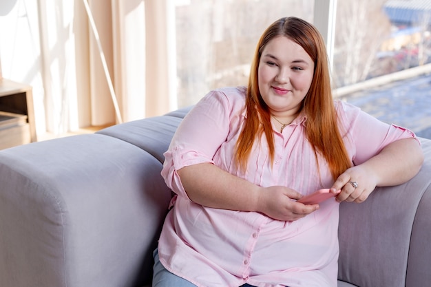 Mujer positiva encantada sosteniendo su teléfono inteligente mientras descansa en el sofá