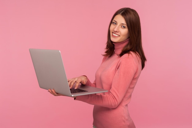 Mujer positiva feliz con cabello castaño en suéter rosa trabajando en la computadora portátil y mirando a la cámara con una sonrisa dentuda disfrutando del trabajo remoto Disparo de estudio interior aislado sobre fondo rosa