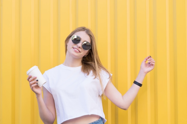 Mujer positiva con gafas y una camiseta blanca con una taza de café en sus manos contra la pared amarilla