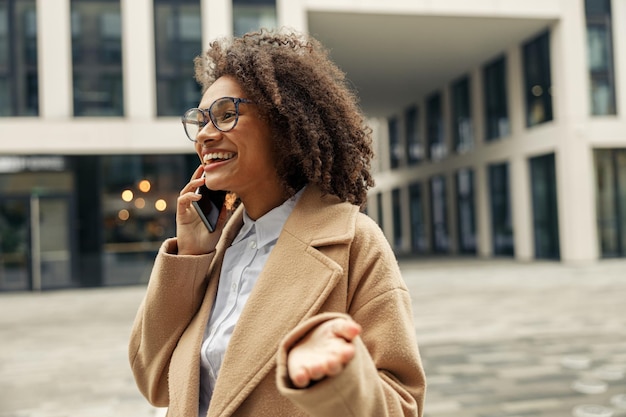 Mujer positiva con gafas hablando por teléfono con amigos durante la caminata por la ciudad