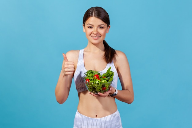 Mujer positiva mostrando los pulgares hacia arriba sosteniendo un tazón grande con ensalada de verduras frescas, manteniendo la dieta de desintoxicación