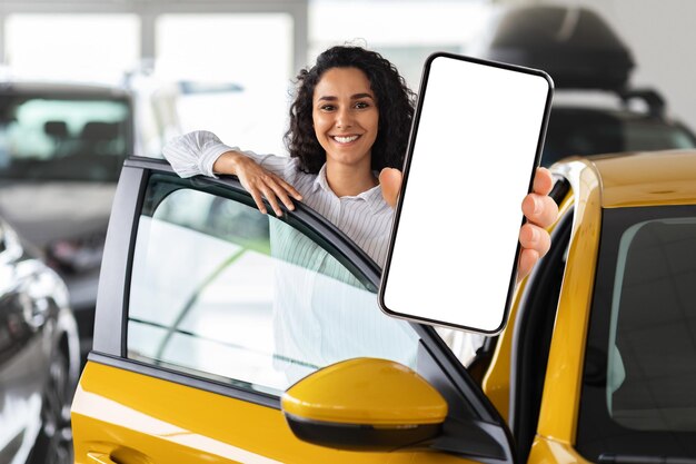 Mujer positiva de Oriente Medio parada junto a un auto mostrando un teléfono inteligente