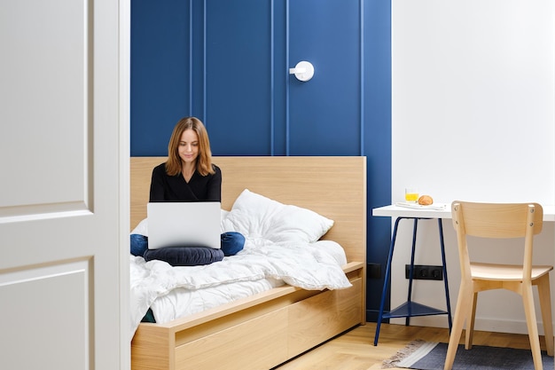 Mujer positiva sentada en la cama en casa y usando una laptop