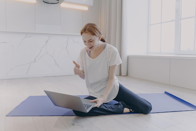 Mujer positiva sentada en el piso con una lección de yoga frente a la computadora portátil Chica muestra los pulgares para entrenar
