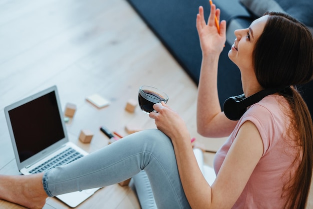 Mujer positiva sentada con una taza de café en el suelo y levantando una mano. Laptop frente a ella