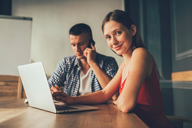 mujer positiva usando una laptop y mirando la cámara mientras se sienta a la mesa con un colega masculino