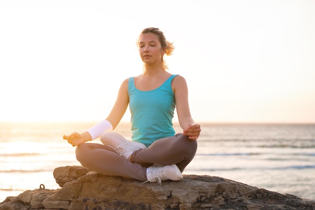 La mujer practica la pose de loto de yoga para la meditación con las vacaciones de verano, la felicidad y la relajación en la playa rocosa. El ejercicio femenino tranquilo con el yoga medita en la playa del océano con el atardecer dorado.