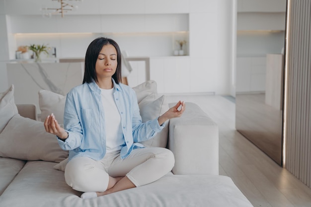 Mujer practica yoga meditando respirando relajándose en posición de loto en el sofá en casa Alivio del estrés
