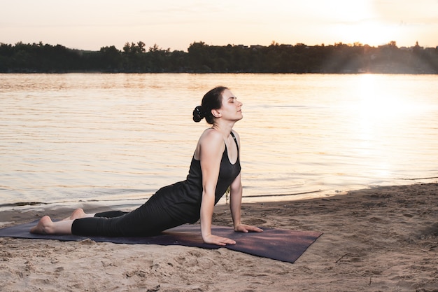 Una mujer practica yoga en la orilla del río en la playa al atardecer.