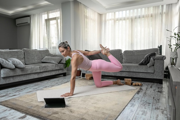 Mujer practicando posturas de yoga que se extienden frente a la computadora portátil de la tableta en casa blogger profesor yoga en línea