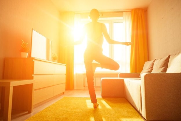 Mujer practicando yoga en casa con ropa violeta de pie en el centro de la sala de estar con grandes ventanales rayos del sol del amanecer