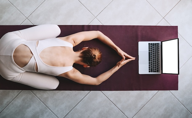 Mujer practicando yoga en una estera de yoga con un portátil Chica está meditando con entrenamiento de vídeo en casa