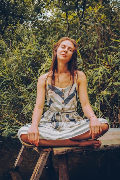 Foto mujer practicando yoga en el muelle junto al río mujer joven con vestido en postura de loto