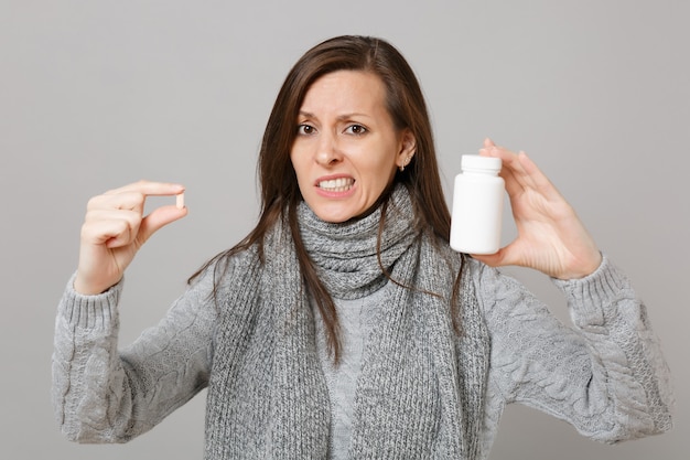 Mujer preocupada en bufanda de suéter gris mantenga tabletas de medicación píldoras de aspirina en botella aislada sobre fondo gris. Estilo de vida saludable enfermedad enferma tratamiento concepto de estación fría. Simulacros de espacio de copia.