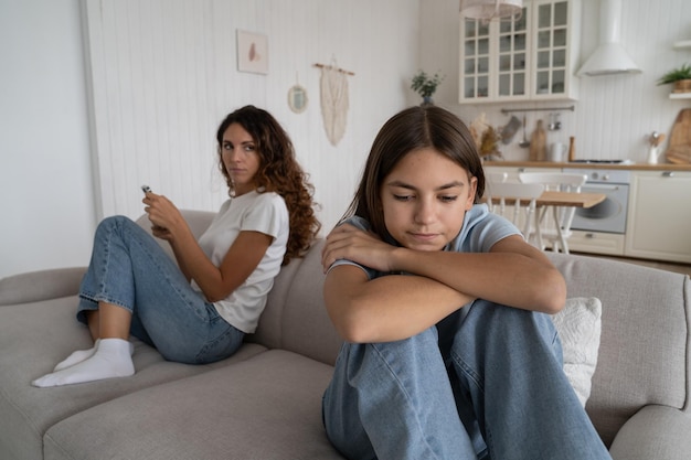 Mujer preocupada casual se sienta en el sofá detrás de una adolescente triste que se inclina sobre las rodillas mira hacia la distancia se siente