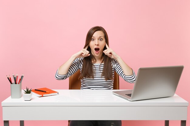 La mujer preocupada no quiere escuchar cubriendo los oídos con los dedos, sentarse y trabajar en el escritorio blanco con una computadora portátil moderna aislada sobre fondo rosa. Concepto de carrera empresarial de logro. Copie el espacio.