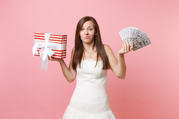 Mujer preocupada en vestido blanco sonriendo con satisfacción extendiendo las manos con un montón de dólares, dinero en efectivo, caja roja con regalo, presente