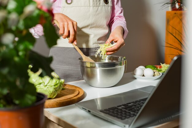 Una mujer prepara una ensalada de verduras en la cocina usando una computadora portátil mirando la receta