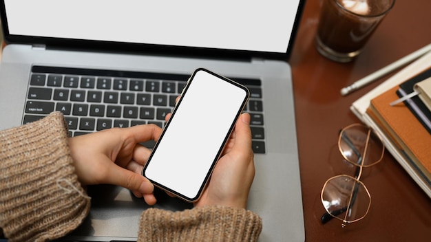 Mujer de primer plano que usa un teléfono inteligente en una computadora portátil en su espacio de trabajo Maqueta de pantalla en blanco del teléfono