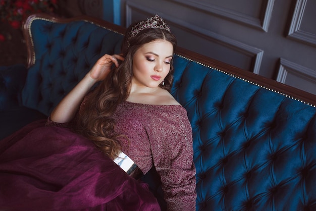 mujer princesa (reina) con un largo vestido de reina púrpura y una corona sentada en el sofá de terciopelo azul