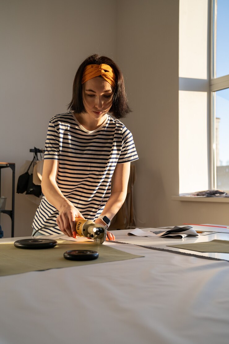 Foto mujer profesional de alcantarillado hacer muestras de ropa en estudio con cortador eléctrico en la mesa en el taller