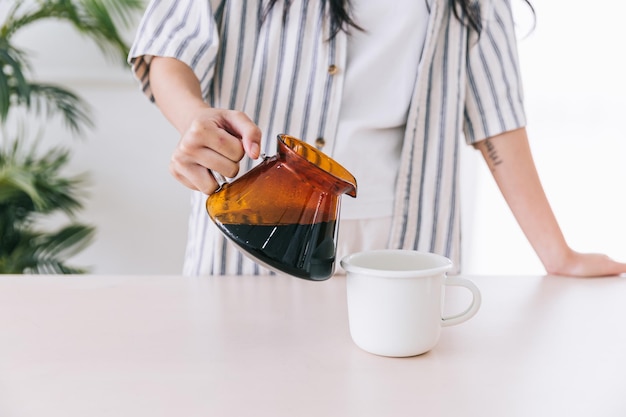 Mujer profesional asiática barista con la mano de pelo largo negro que vierte café de goteo recién hecho de un gotero de vidrio en una taza en casa Preparando el método alternativo del café