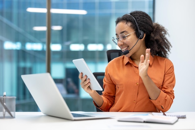 Una mujer profesional sonriente se involucra a través de una tableta y una computadora portátil con auriculares para la comunicación