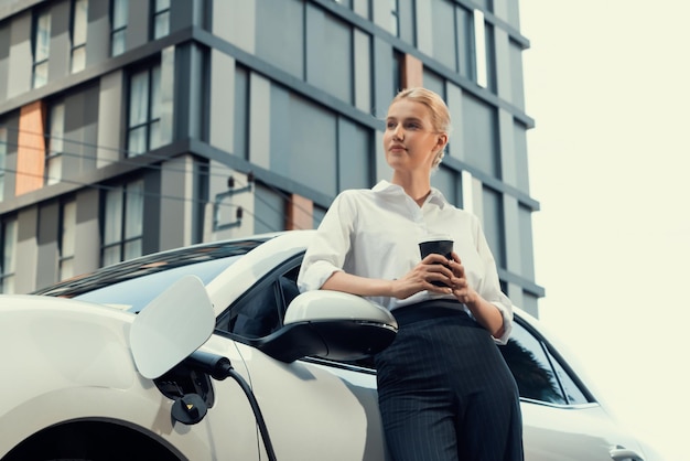 Mujer progresista con café mientras carga un coche EV con edificios residenciales