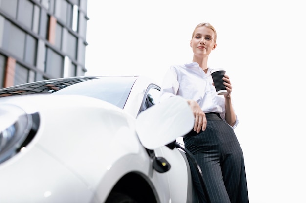 Mujer progresista con café mientras carga un coche EV con edificios residenciales