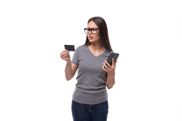 Foto mujer promotora joven con gafas vestida con una camiseta gris sostiene una tarjeta de plástico y un teléfono inteligente en un