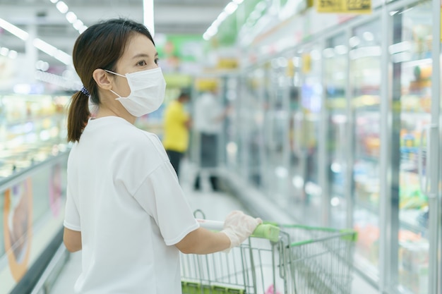 La mujer se protege de la infección con la máscara quirúrgica y los guantes, con un carrito de compras para comprar en el supermercado después de la pandemia de coronavirus.