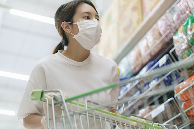 La mujer se protege de la infección con la máscara quirúrgica y los guantes, con un carrito de compras para comprar en el supermercado después de la pandemia de coronavirus.