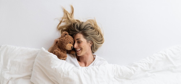 mujer que se acurruca con su oso de peluche durmiendo en una cama blanca y acostada bajo una manta, Vista superior, espacio de copia para su texto individual.