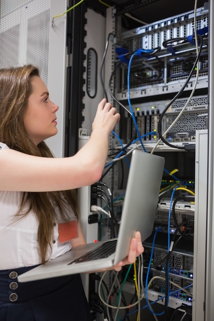 Mujer que ajusta los alambres del servidor y que sostiene la computadora portátil