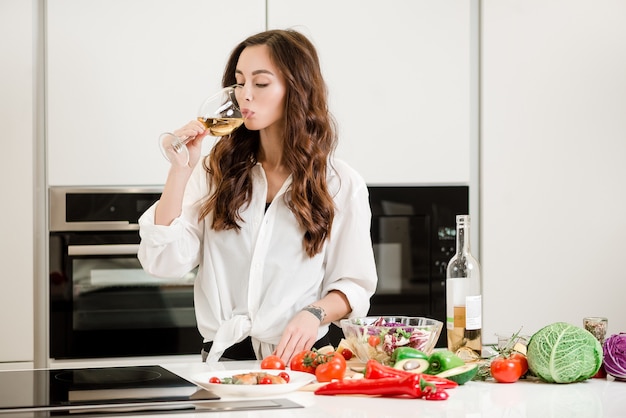 Mujer que bebe un vaso de vino blanco en la cocina mientras cocina ensalada