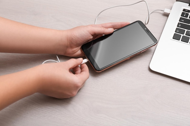 Mujer que conecta el teléfono inteligente con el cable de carga a la computadora portátil en el primer plano de la mesa gris claro Espacio para texto