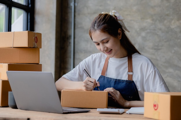 Una mujer que dirige un negocio de comercio electrónico está escribiendo una lista de clientes en papel antes de enviárselos. Dirige un negocio de comercio electrónico en sitios web y redes sociales. Concepto de venta de productos en línea.