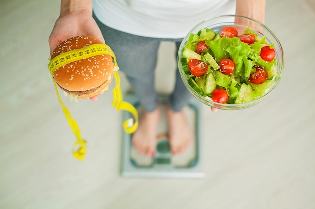 Foto mujer que mide el peso corporal en la balanza con hamburguesa y ensalada.